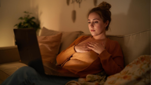 Frau Anfang 30 sitzt abends mit Laptop auf dem Schoß auf dem Sofa, hält eine Hand auf dem Herzbereich und schaut in die Kamera, während sie an ihrer Bindungsangst arbeitet und Wege sucht, ihre Bindungsangst zu überwinden.